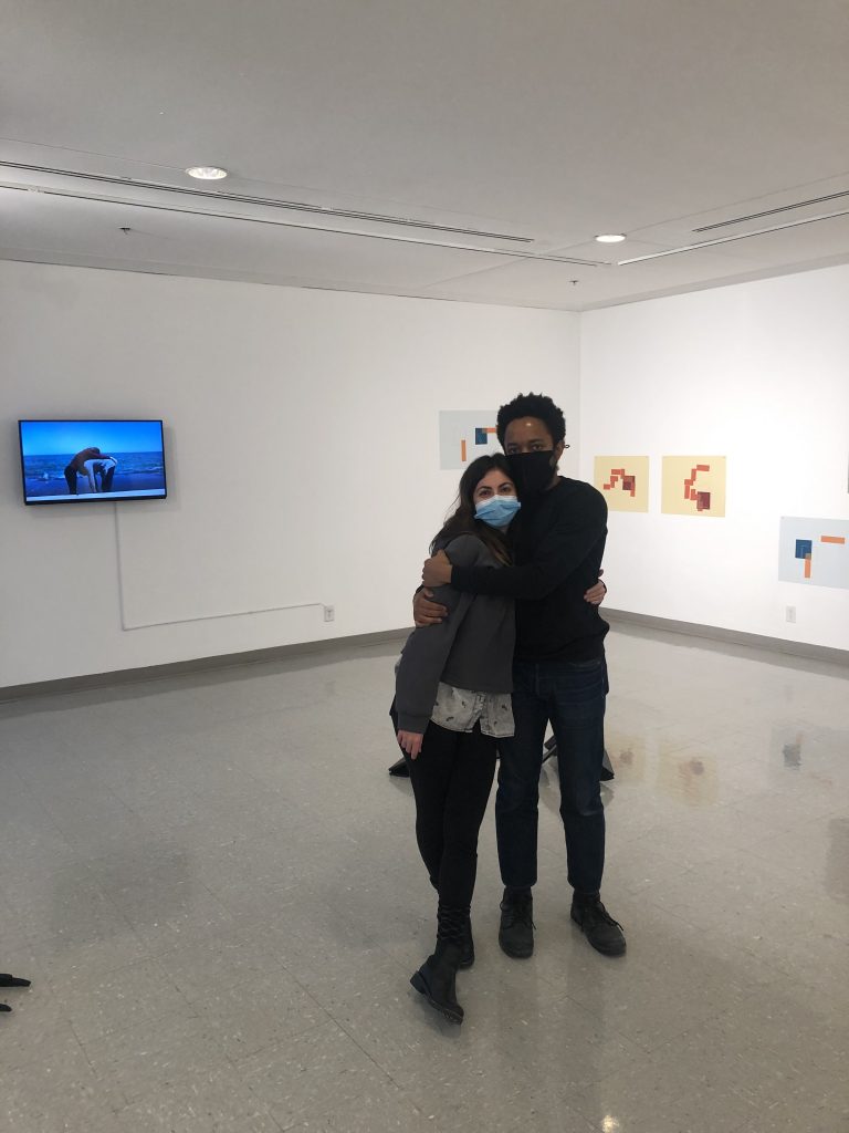 Image: Shir and Max stand in the middle of the gallery at University of Illinois Springfield. Several of their works are visible in the background, including the video work How to Make Windows for the Horizon. Photo courtesy of the artists.