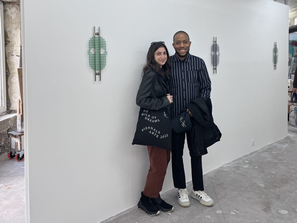 Image: Max and Shir are seen together in an exhibition space, standing in front of a wall where three of Max's works are mounted. Photo courtesy of the artists.