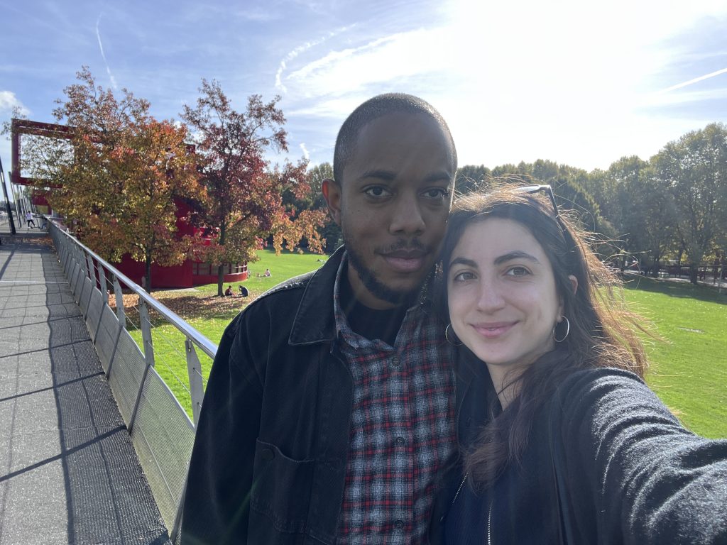 Image: Max and Shir standing outside on a fall day, the sun blazing in the background through a blue sky. Photo courtesy of the artists.