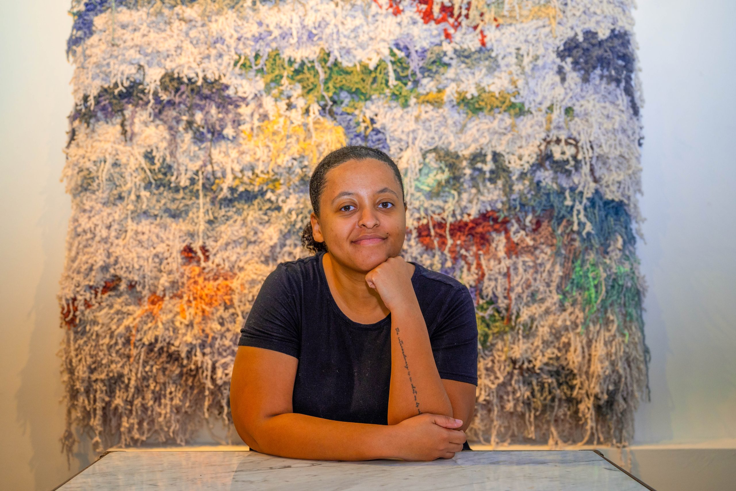Image: Bryana Bibbs poses in front of her woven artwork Numb, created in 2023. The woven artwork is approximately six feet tall and six and a half feet wide. The color of the woven yarns is predominately white with speckles of other colors including hues of blue, purple, red, brown, and yellow. Photography by Tonal Simmons.