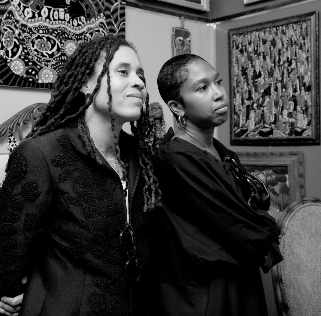 Image: A black-and-white image of Cierra and zakkiyyah standing in a room where the walls are covered with artworks, they're both looking beyond the frame. Photo courtesy of the artists.