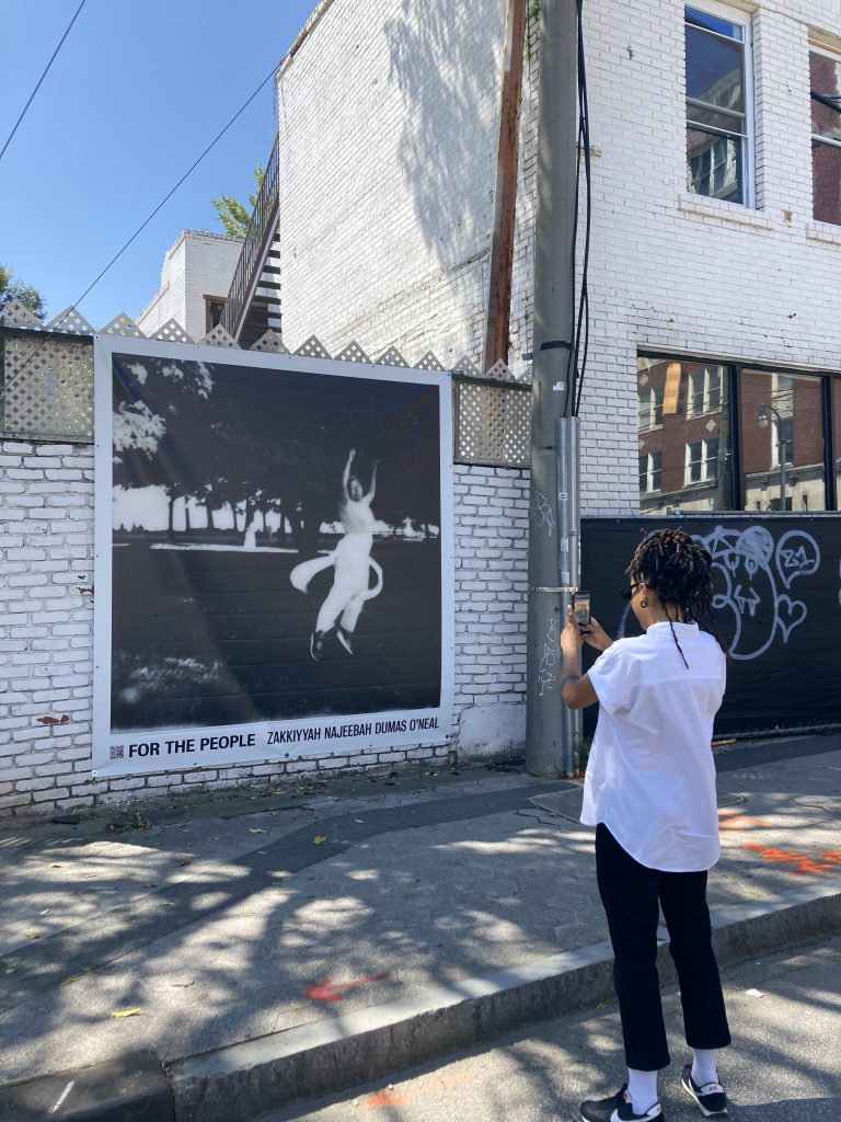 Image: Cierra is seen snapping a photo in front of a large outdoor sign for zakkiyyah's "For the People," which is posted up on a white brick wall. Photo courtesy of the artists.
