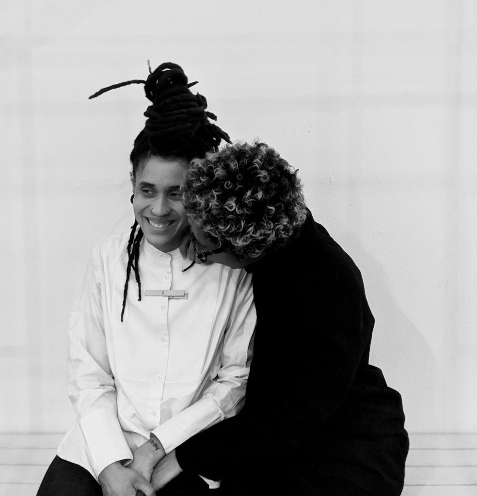 Image: A black-and-white photo of Cierra and zakkiyyah sitting together, hugged up against a white wall. Photos courtesy of the artists.