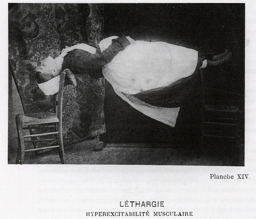 Image: A black and white photograph of a woman in a traditional French maid uniform impossibly floating, suspended, between two wooden chairs.