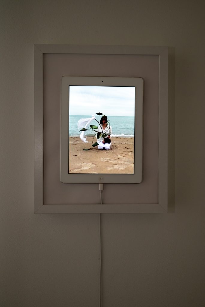 Image: Quiet Chaos, Laleh Motlagh. A framed iPad features an image of Motlagh kneeling on a beach. Behind her is water. Image courtesy of the artist.