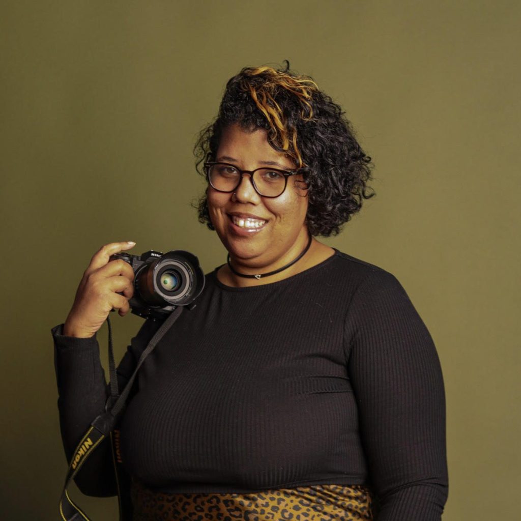 Image: A headshot of photographer Tonal Simmons holding a camera and smiling at the viewer.