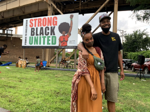 Image: Janell and Joe stand outside in front of a large sign that says "Strong, Black, United." Photo courtesy of the artists.