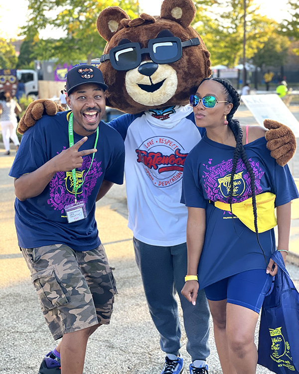 Image: In an outdoor scene, Joe and Janell pose on either side of a person in a bear costume, wearing matching tees. Photo courtesy of the artists.