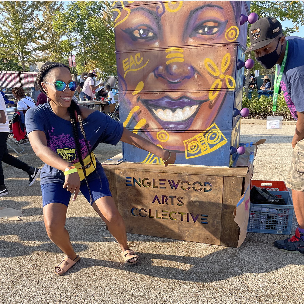 Image: Janelle and Joe pose playfully on either side of a sculpture. The piece has a smiling face is painted on the front and it stands on a platform that has the words Englewood Arts Collective  across the front. Image courtesy of the artists. 