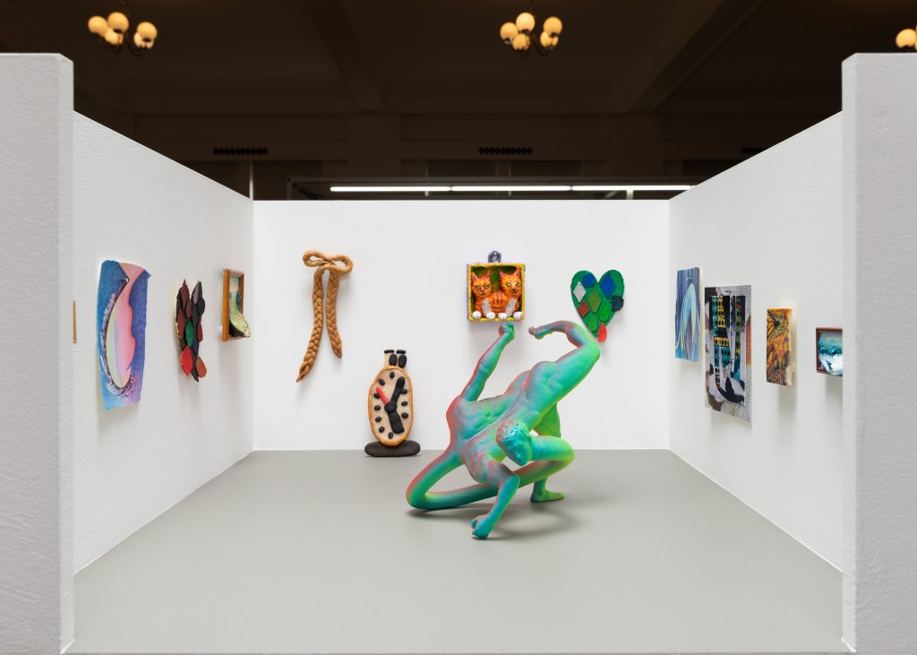 Image: An installation view of Good Naked's booth at Barely Fair, featuring a variety of different 2D and 3D piece by various artists. In the center is a sculpture of a green, twisted human-like body. Photo by roland miller.