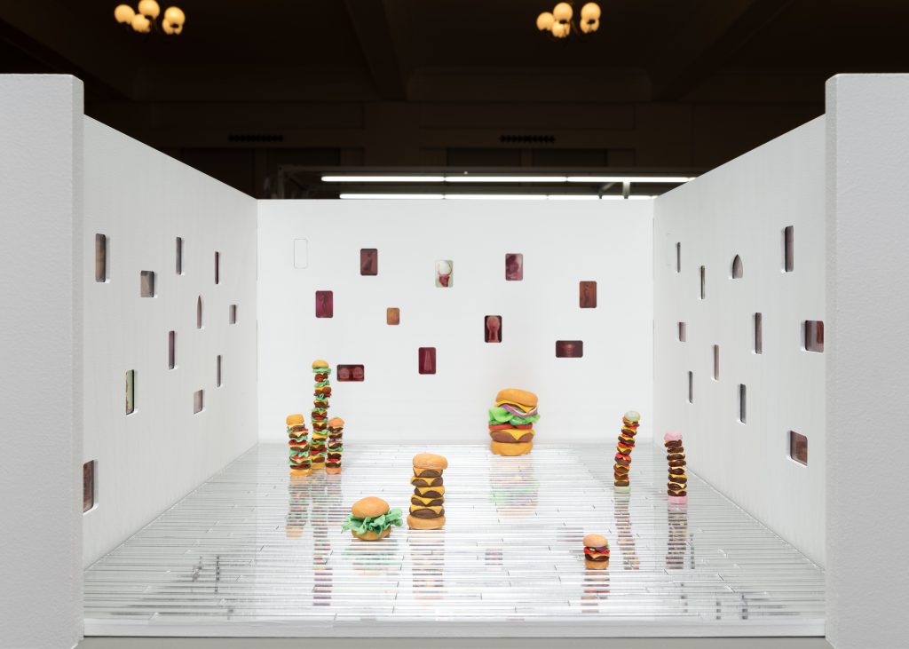 Image: An installation view of Flyweight's booth at Barely Fair, featuring the work of Yael Eban and Jeni Emery. Tiny images are sprinkled across three white walls, and stacks of cheeseburgers sit on a silver, reflective floor. Photo by roland miller.