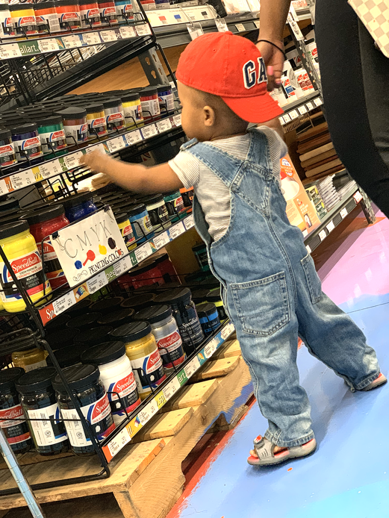 Image: A scene from the art store with their son, who is wearing overalls and a red cap, standing in front of art supplies and reaching for a jar. Photos courtesy of the artists.