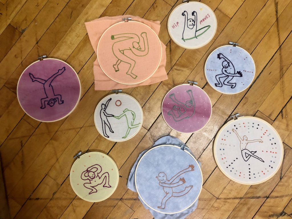Image: Nine embroidery hoops of multiple colors are seen gathered on a hard wood floor. Each hoop has a character in a pose or in the middle of a movement or dance. Photo courtesy of the artist. 