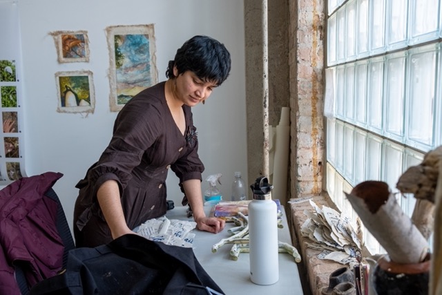 Image: Artist Kushala Vora in her studio in Chicago, Illinois. She stands over her studio desk and organizes ceramic pieces. Photo courtesy of Saadia Pervaiz.
