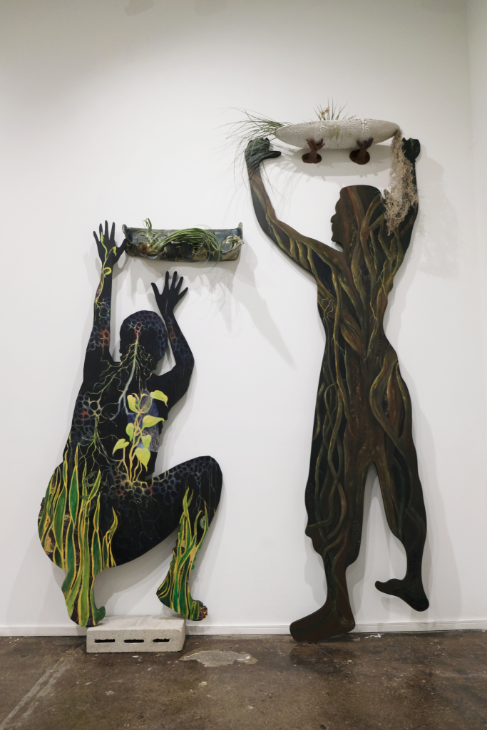 Image: Two of Ladsariya’s silhouette pieces at her BOLT installation at the Chicago Artists Coalition. Two painted silhouettes of the artist are posed in a kneeling and standing position, both with arms raised, holding vessels for air plants and aloe. Photo by Saadia Pervaiz.