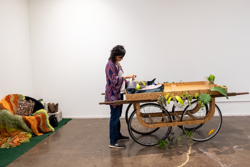 Image: Artist Tulika Ladsariya stands at her haath-gadi in the BOLT gallery space planting a propagated plant to share with visitors. Photo by Saadia Pervaiz.