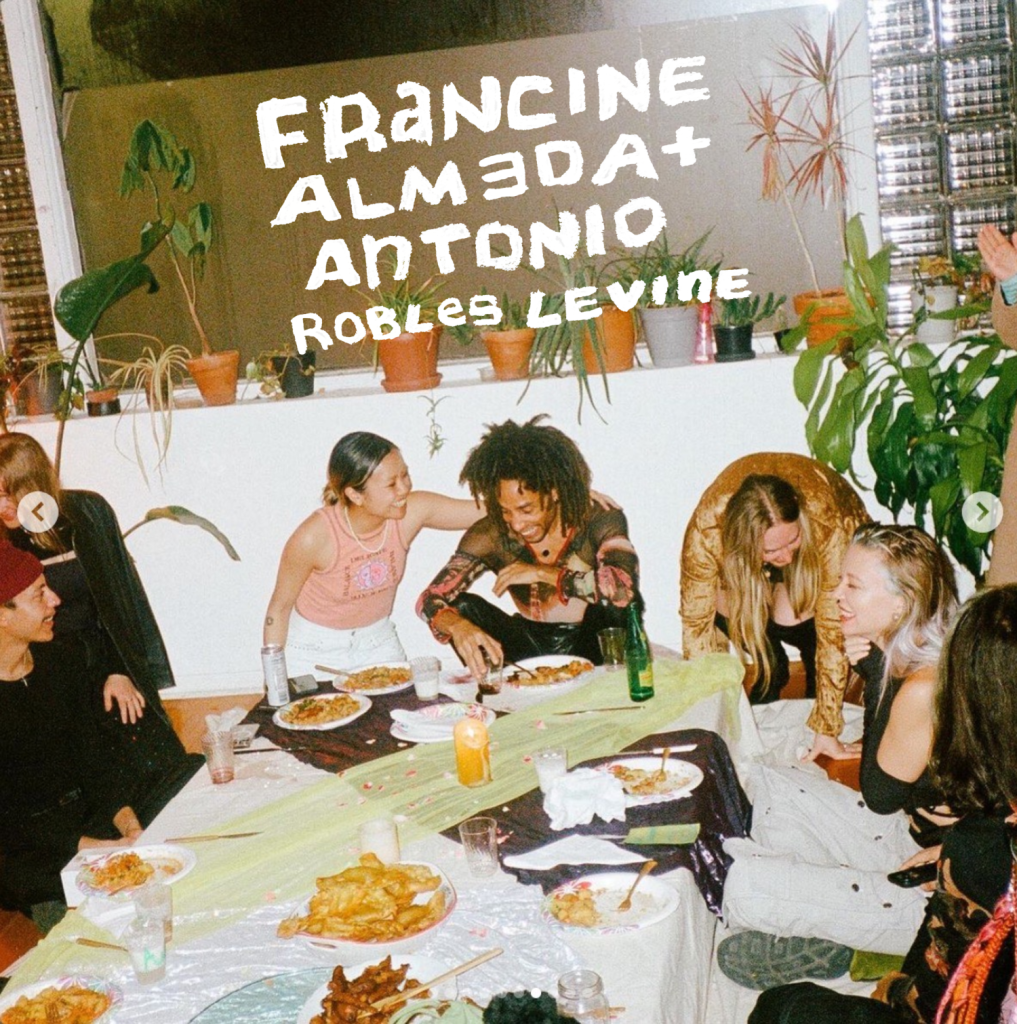 Image: Francine and Antonio sit at the end of a low dinner table surrounded by friends. Francine’s arm is wrapped around Antonio as they both are laughing. Their names can be seen going across the top of the photo. Photo by Steven Piper.