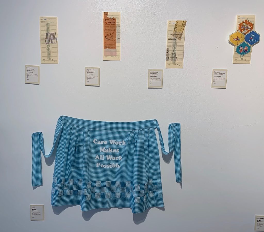 Image: Installation view, Ghost Prints and Shadow Work, 2022. The Compassion Factory, Brookfield, IL. Vintage, embroidered apron with vinyl lettering. Four "Time Cards for Mothers," are attached to a white wall in an art gallery. Below the time cards, there’s a vintage, handmade apron with added white, vinyl, text that says, “Care Work Makes All Work Possible.” The apron is blue and white gingham and is probably from the 1960s. Courtesy of the artist.