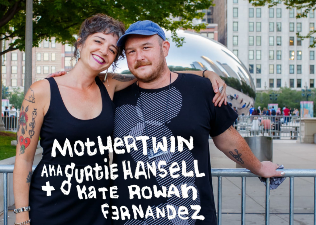 Image: kate and gurtie stand along a fence in Chicago's Grant Park, both wearing black t-shirts. The Bean (a.k.a. Cloud Gate) can be seen in the background. Their names can be seen going across their torsos. Photo by Sarah Elizabeth Larson, courtesy of the artists.