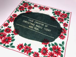 Image: Your Creator of Christmas Magic Day was Mom, 2022. Vintage, commercially printed handkerchief, printed with light reactive dye to add text (photogram). "Your Creator of Christmas Magic Today was Mom," vintage commercially printed handkerchief printed with light reactive dye by Elaine Luther. Poinsettias are all around the edge of the handkerchief. In the center, a green oval has been painted on and the text is in white. Courtesy of the artist.