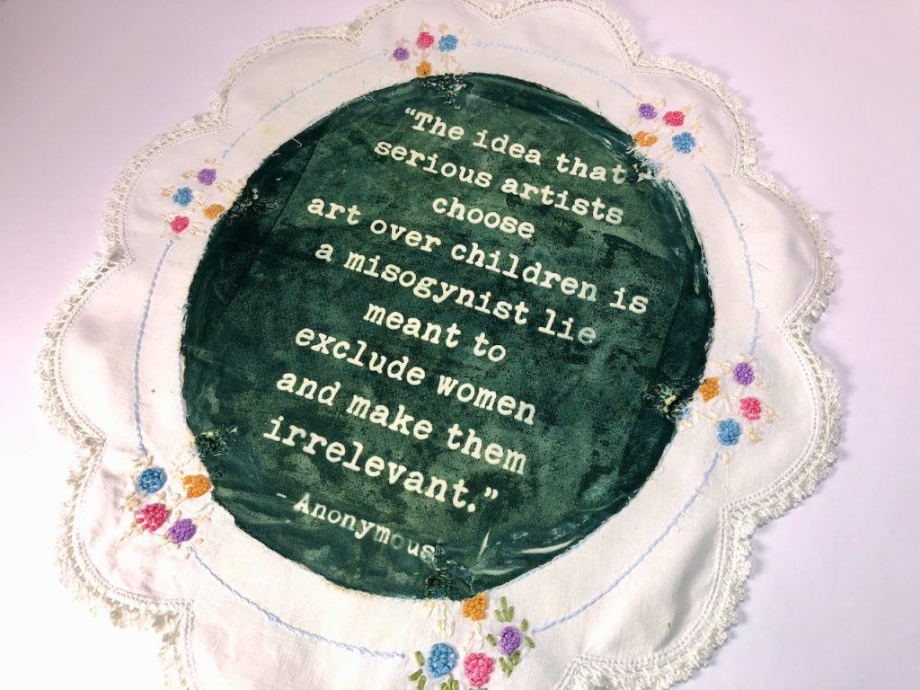 Image: Elaine Luther, Misogynistic Lie, 2022. The text says: “The idea that serious artists choose are over children is a misogynistic lie meant to exclude women and make them irrelevant. - Anonymous.”  A white oval-shaped doily has colorful floral embroidery around the edges. The center has been painted green with white text. 