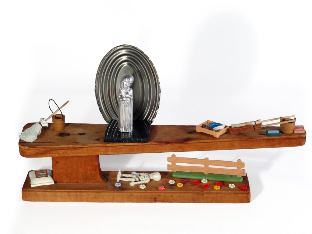 Image: Elaine Luther, Our Lady of Perpetual Housework, 2014. 18 x 9.5 x 3.5 in. Found ironing board for sleeves, silver-leafed saint, dollhouse miniatures, PlayMobil skeleton, fence, flowers, mice, and household objects; sardine can lid and marble tile. An old wood ironing board that was used for ironing sleeves is the base for this sculpture.  Slightly off center on the top level of this two-level piece of wood is a small square of black marble, on top of which stands a plastic saint figure, which has been silver leafed.  Behind the saint figure is a large oval lid from a metal food can. To each side of the saint, there are miniature wooden buckets, a mop, a broom, packets of cleaning products, and plastic mice. On the lower level of the piece of wood, there’s a bag of gardening supplies, a piece of plastic fencing, tiny plastic flowers, and a plastic toy skeleton. Courtesy of the artist.