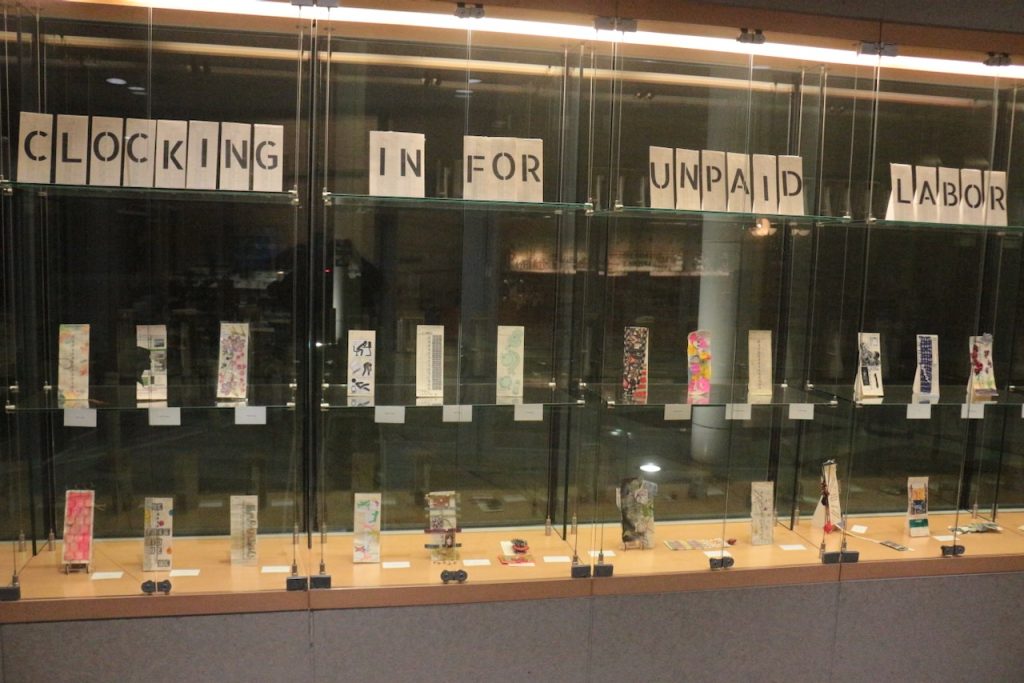 Image: Installation View of Clocking in for Unpaid Labor by Elaine Luther at Orland Park Public Library, Orland Park, IL, 2023. Multiple artists,** Approximately 55 time cards on custom-made easels, plus collaged time cards with titles, and a vintage time clock. Courtesy of the artist.