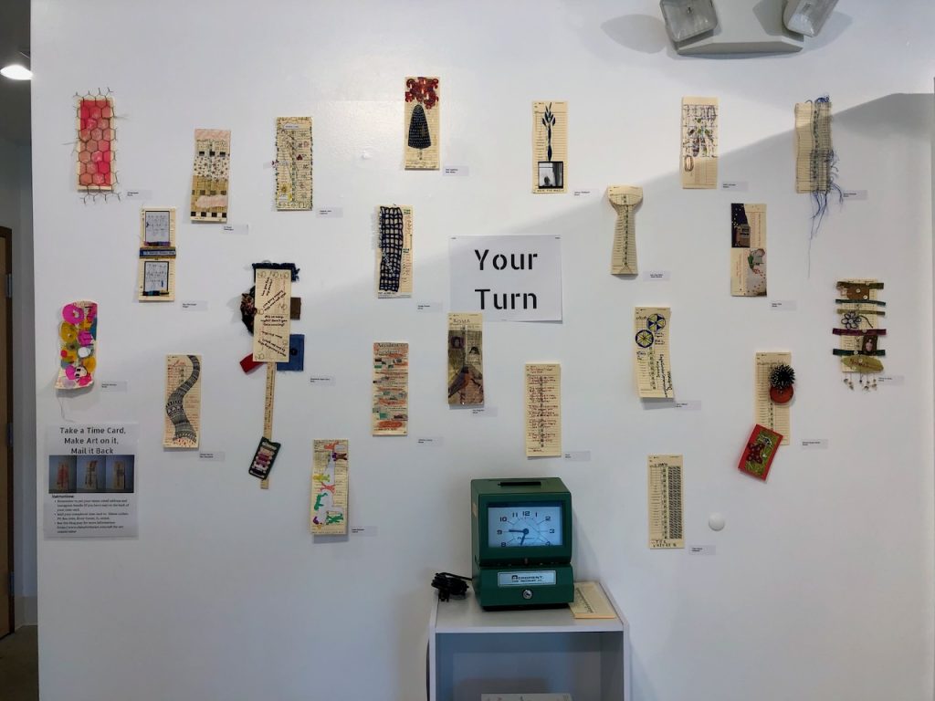 Image: Installation view, Ghost Prints and Shadow Work, 2022. The Compassion Factory, Brookfield, IL. Vintage, embroidered apron with vinyl lettering. Four "Time Cards for Mothers," are attached to a white wall in an art gallery. Below the time cards there’s a vintage, handmade apron with added white, vinyl, text that says, “Care Work Makes All Work Possible.” The apron is blue and white gingham and is probably from the 1960s. Courtesy of the artist.