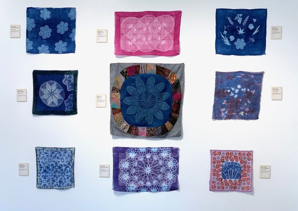 Left Image: Installation View of Ghost Prints and Shadow Work, 2022 by Elaine Luther. The Compassion Factory, Brookfield, IL. Photograms of vintage doilies printed with light reactive dye onto vintage handkerchiefs, placemats, napkins, and quilt top fragments. Vintage textiles, light reactive dye, and colored pencils. Two Gallery Walls at Ghost Prints and Shadow Work by Elaine Luther. Many printed handkerchiefs, napkins, and placemats. Each one is a photogram.  The vintage linens mostly began as white and are now purple, blue, or magenta.  In the center is a quilt fragment of gray fabric with pieced quilt elements and a photogram in the center, created by making a print (photogram) using a vintage doily. Right Image: Ghost Runner 2, 2022. The runner is white with elaborate blue embroidery around the edges. In the center are three circles in blue, each one a different photogram. The center circle is a print of two ginkgo leaves. On either side of that are two larger circles, each with a print (photogram) of a vintage doily with elaborate designs. Courtesy of the artist.