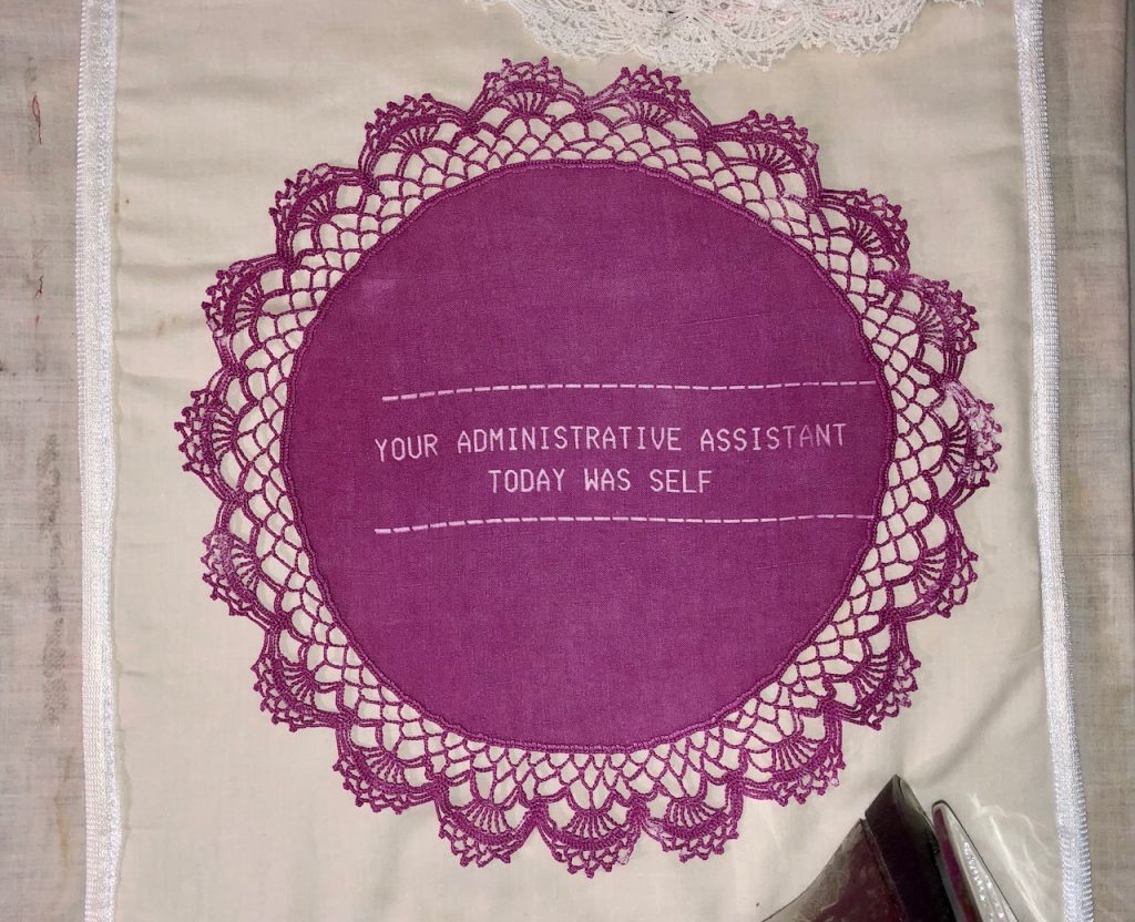 Image: Elaine Luther, Your Administrative Assistant Today was Self, 2022. The magenta doily says "YOUR ADMINISTRATIVE ASSISTANT TODAY WAS SELF,” The doily has elaborate crocheted edgework that extends beyond the doily itself by about two inches. Both images courtesy of the artist.