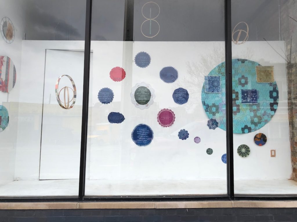 Image: Installation View of Balance & Tension, 2023 by Elaine Luther. Ignition Project Studios, Chicago. Doilies with quotes on them and additional works hang on a white wall, which can be seen through a storefront window. Courtesy of the artist.