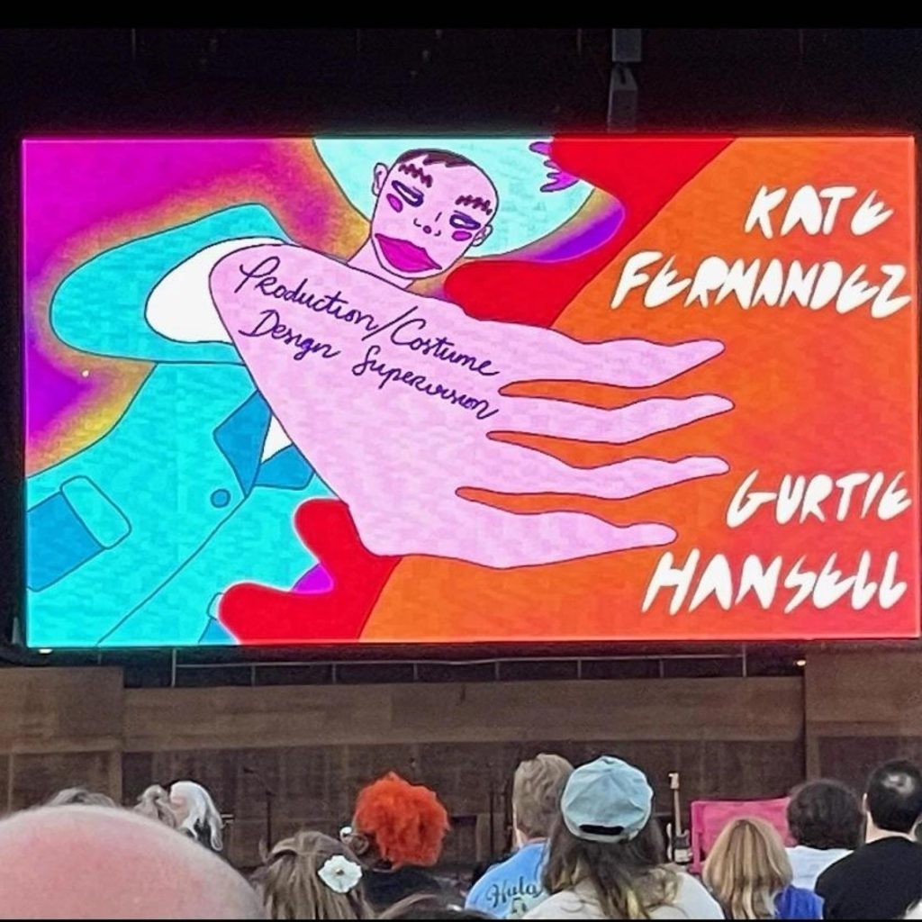 Image: An animation is projected on a giant screen in front of an audience. The animation shows a figure wearing a bright blue suit and hat, their hand in front of them taking up most of the screen, inscribed with the words "production/costume design supervisor." The words "KATE FERNANDEZ" and "GURTIE HANSELL" appear in white text to the right. Animation by Zoe Pham for the Andrew in Anotherland feature length starring Andrew Sa, directed by Jesse Morgan Young. Photo by kate rowan fernandez. 