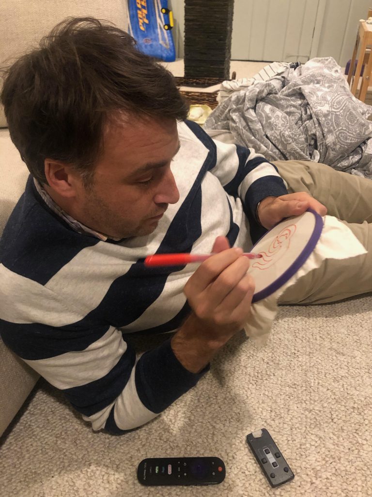 Image: Scott is  seen laying down, embroidery hoop and a bright red parker in hand, focused on drawing a sketch onto a small piece of fabric. Photo courtesy of the artists.