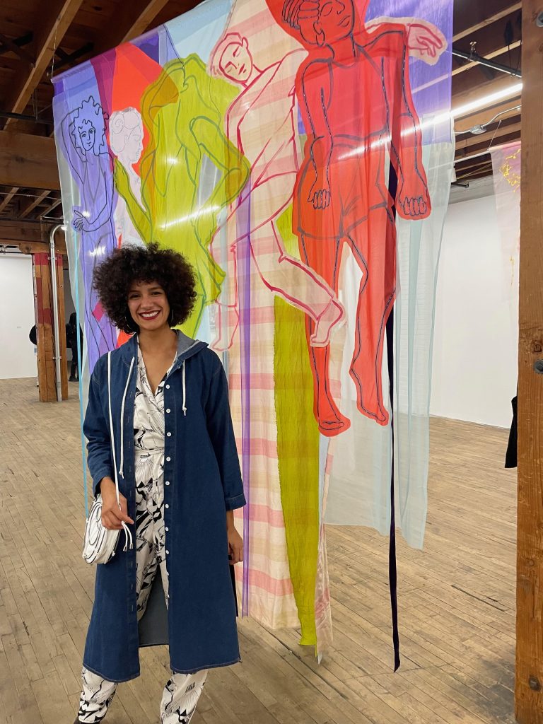 Image: Alexandra is seen in an exhibition space with a large colorful textile piece hanging down from the ceiling. Photo courtesy of the artists.