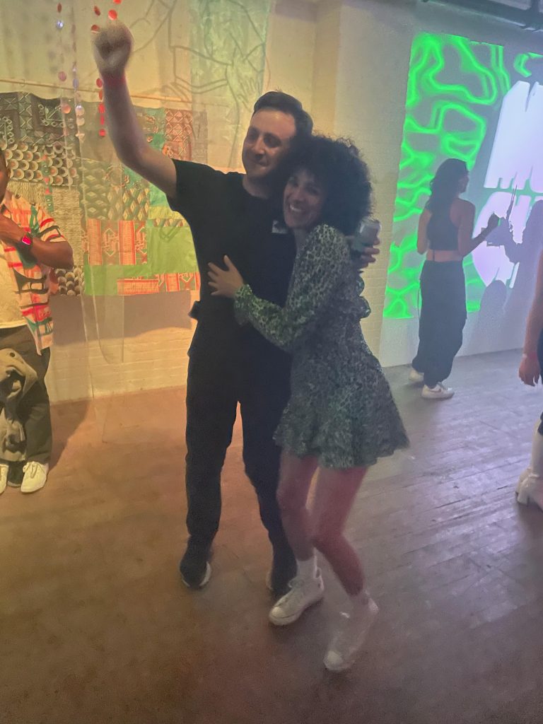 Image: Alexandra and Scott hug and pose for the camera at an event and exhibition space. Other attendees and multiple works can be seen in the background. Photo courtesy of the artists.