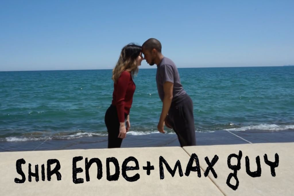 Image: Shir and Max stand facing one another, leaning and touching foreheads against a clear blue sky and Lake Michigan. The image has their names across the bottom. Still courtesy of the artists.