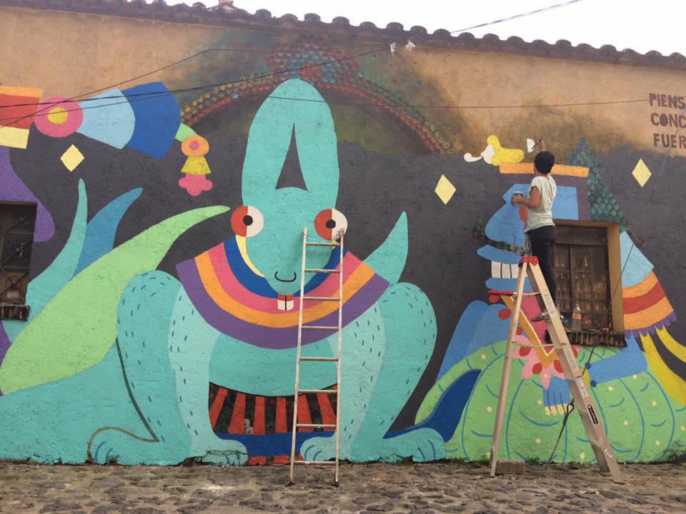 Imagen: Una foto de Cecilia Beaven pintando un mural en México. Puedes ver a Cecilia parada en una escalera, de espaldas al lente. Foto cortesía de la artista. // Image: A photo of Cecilia Beaven painting a mural in Mexico. You can see Cecilia standing on a ladder, back facing the lens. Photo courtesy of Cecilia Beaven.