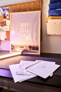 Image: The desktop of Gabriel Cuillier and Charli Andrews' studio. A stack of papers from the script of Buy Sell Trade are in the foreground. In the background is a framed poster that says "Buy Sell Trade."