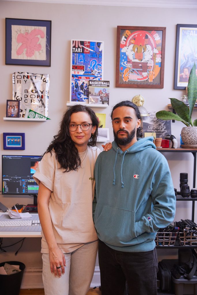 The couple Charli Andrews, and Gabe Cuillier, take a portrait in their home studio. The walls of their stuiod are cream colored and filled with paintings, shelves, and plants. There is a desk with a computer behind them showing film editing software in use. Charlie is the left with her hand on Gabe's shoulder. Charlie is wearing light tan colored shirt and pants, and person on the right, Gabe is wearing a light blue hoodie and black jeans.