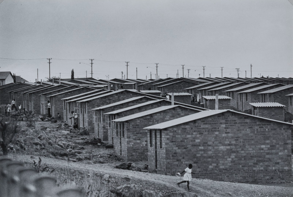 Image: A black and white image of rows of one-story brick houses with a young girl in a light dress running down a dirt road. Ernest Cole, From “House of Bondage,” 1960s, Gelatin silver print. Smart Museum of Art, The University of Chicago, Gift of the Estate of Lester and Betty Guttman, 2014.224. © Ernest Cole / Magnum Photo.