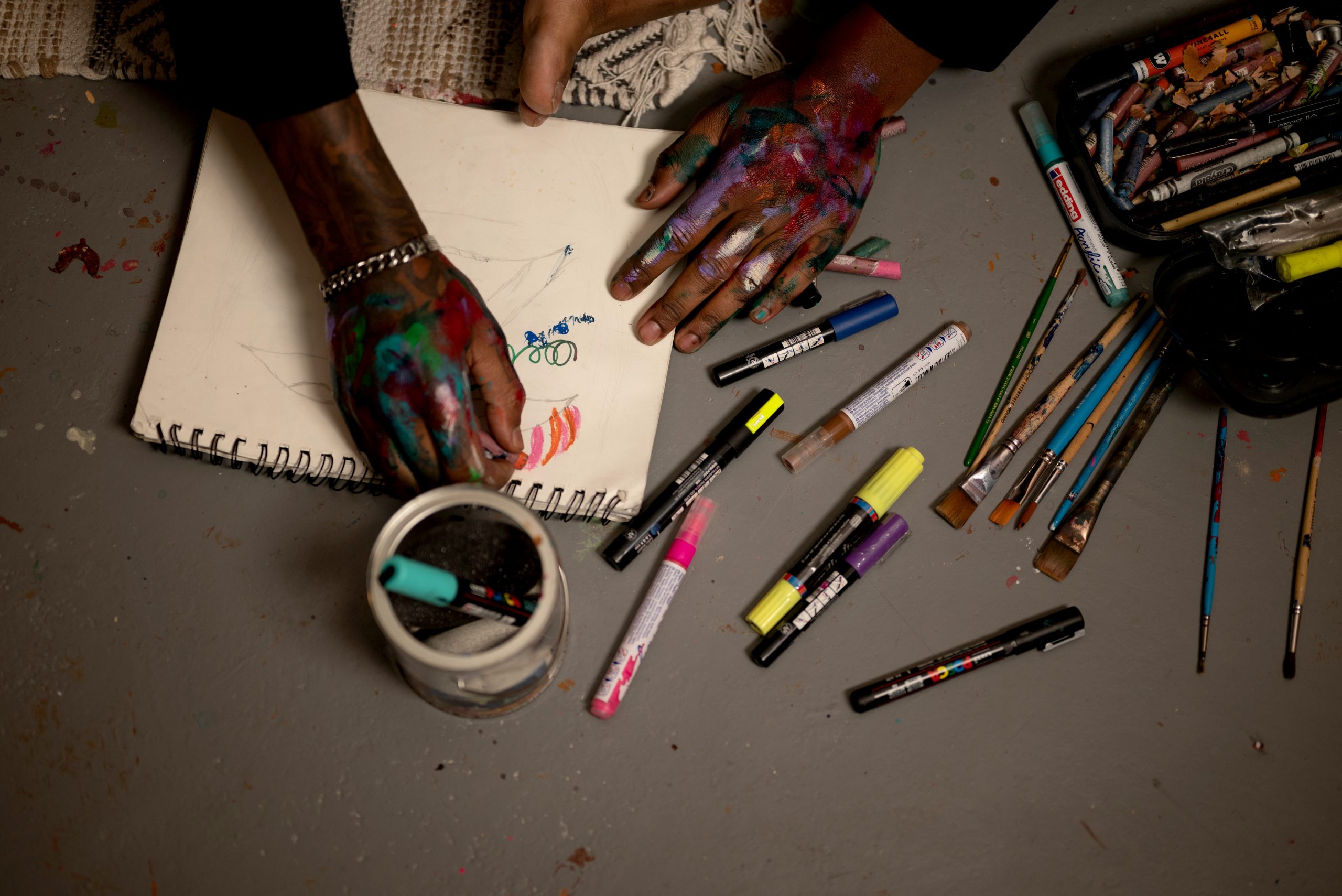 Image: top down view of a floor. Art supplies, markers, and pens scattered across the table. On a piece of paper, a pair of hands drawing. There are swatches of paint on the hands. Photo by Tonal Simmons.
