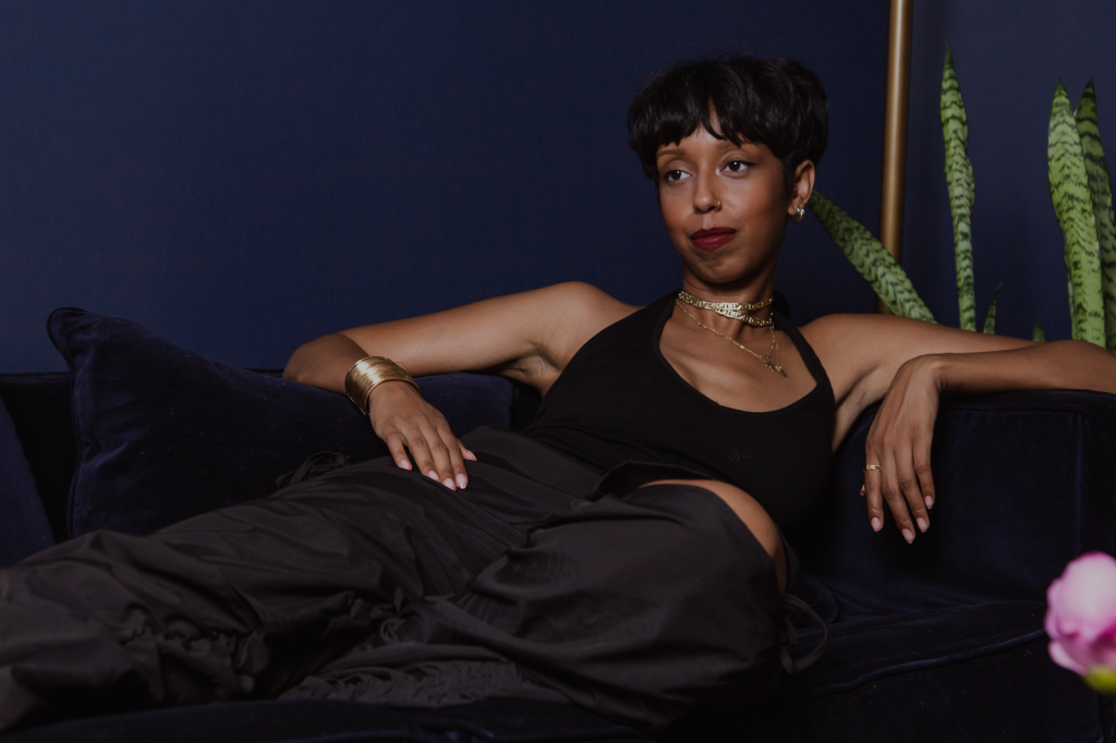Image: The portrait features Barédu sitting on a dark blue couch in front of a dark blue wall. Behind her, there is a gold lamp stand, a snake plant, and a pink flower in the lower right-hand corner. She has on a black top and black pants and is wearing gold necklaces, bracelets, and earrings. Image captured and edited by Joshua Johnson.