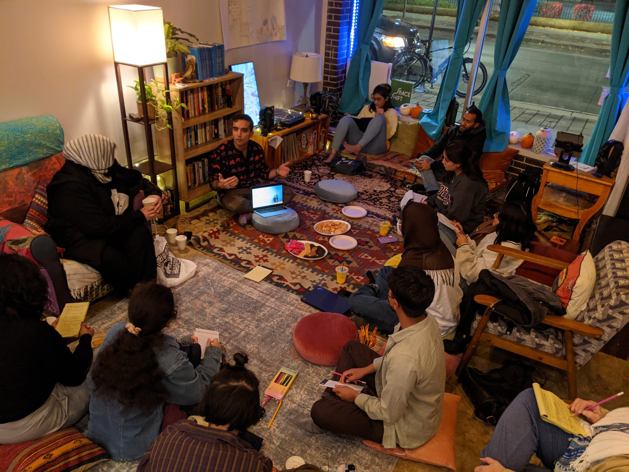 Image: Community members gather in Starlight’s storefront space for a storytelling workshop. People with notepads and pencils sit on various rugs in an ambiently-lit storefront. There are snacks and drinks on the rugs in the center of the room. Courtesy of the SpaceShift Collective.