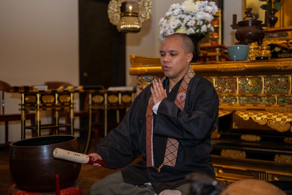 Image: Brown seated in front of a golden shrine. They hold one hand up towards their face and the other is holding a mallet up against a bowl. Photo by Tonal Simmons.