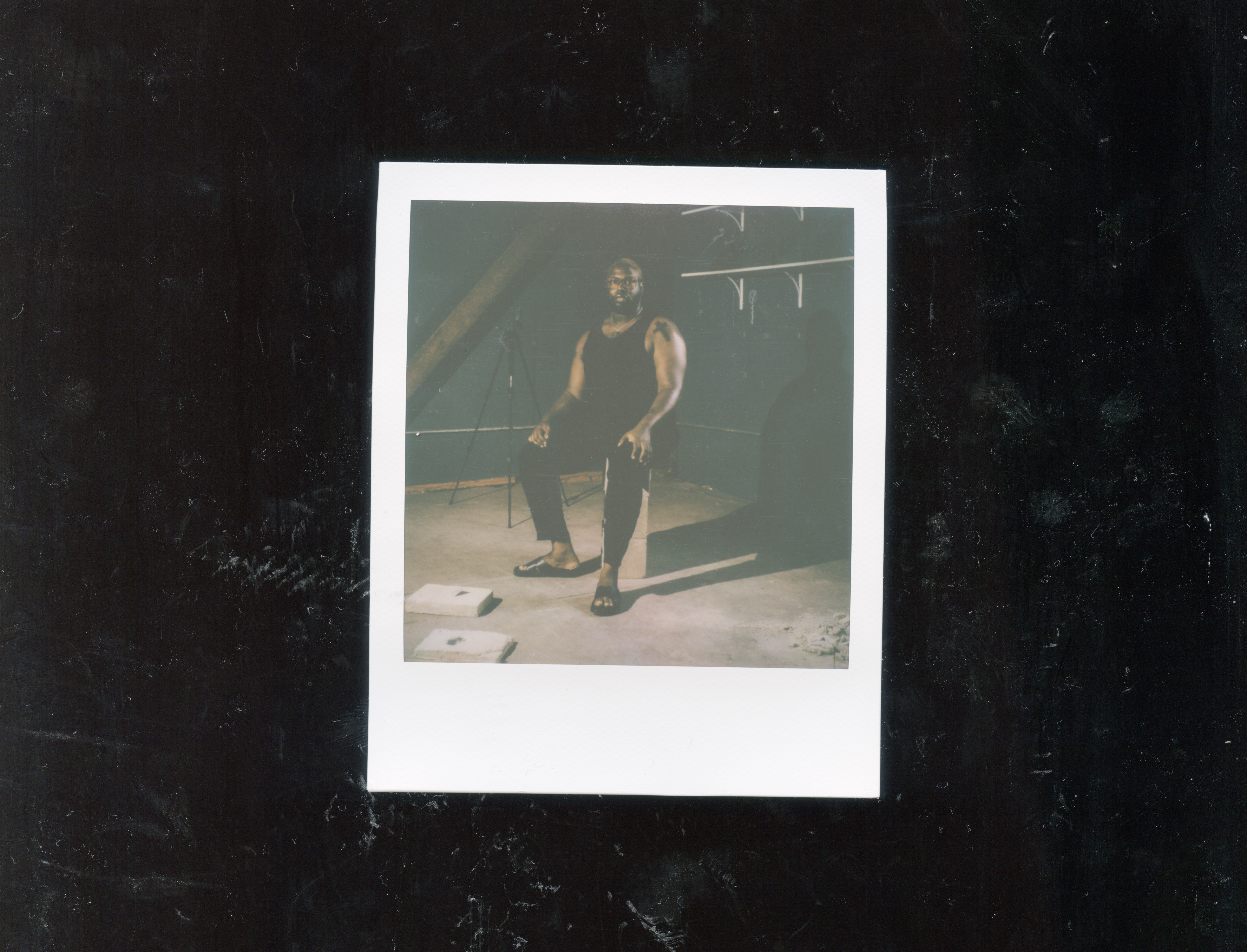 Image: In this scanned self-portrait there is a Polaroid of Shabez Jamal in their studio, wearing all black and seated on a stack of concrete bricks. There are concrete artworks at their feet and in the background is a dark blue wall and shelves. The Polaroid is scanned and placed within the center of the image, floating against a dark black, textured background. Photo by Shabez Jamal.