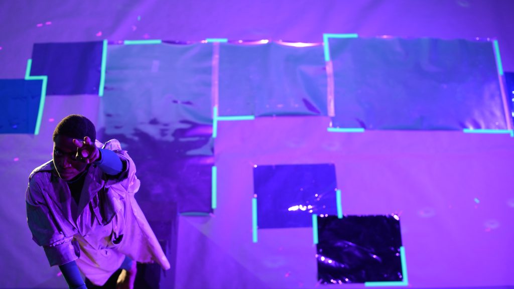 Image: A performer gazes at the audience while pointing two fingers at them. The stage is lit with a purple light making colors appear neon. The picture was taken at Experimental Media and Performing Arts Center by Will Rawls. Courtesy of the Will Rawls and MCA.