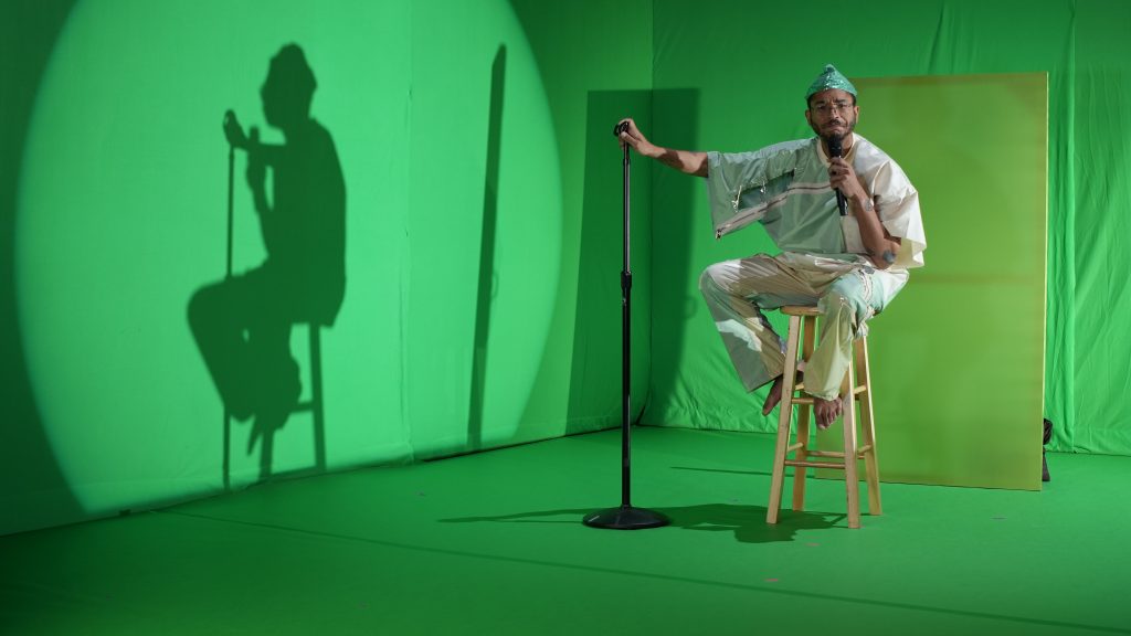 Image: In the spotlight, performer Jeremy Toussaint-Baptiste sits on a stool with a microphone in hand. The stage has a green floor and a green backdrop. Also on stage is a large blank canvas with a different performer hiding behind it. The picture was taken at Experimental Media and Performing Arts Center by Will Rawls. Courtesy of the Will Rawls and MCA.