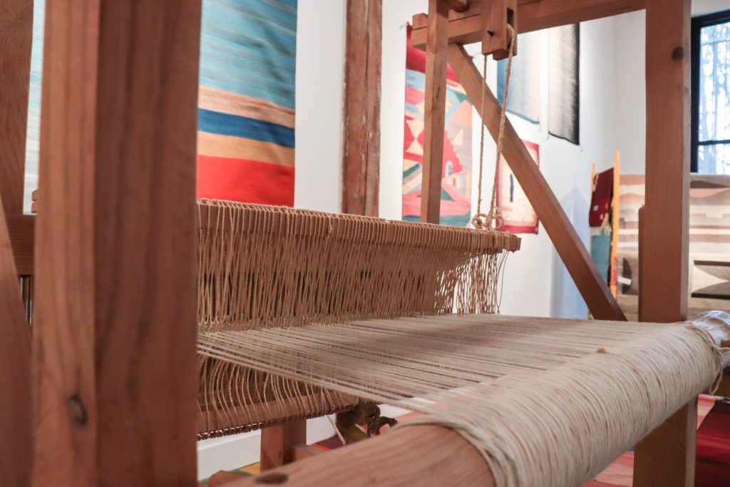 Image: Loom machine inside MANO Gallery, 2023. A wooden loom can be seen with several colorful textile pieces hanging on a wall in the background. Photo by Livy Snyder.