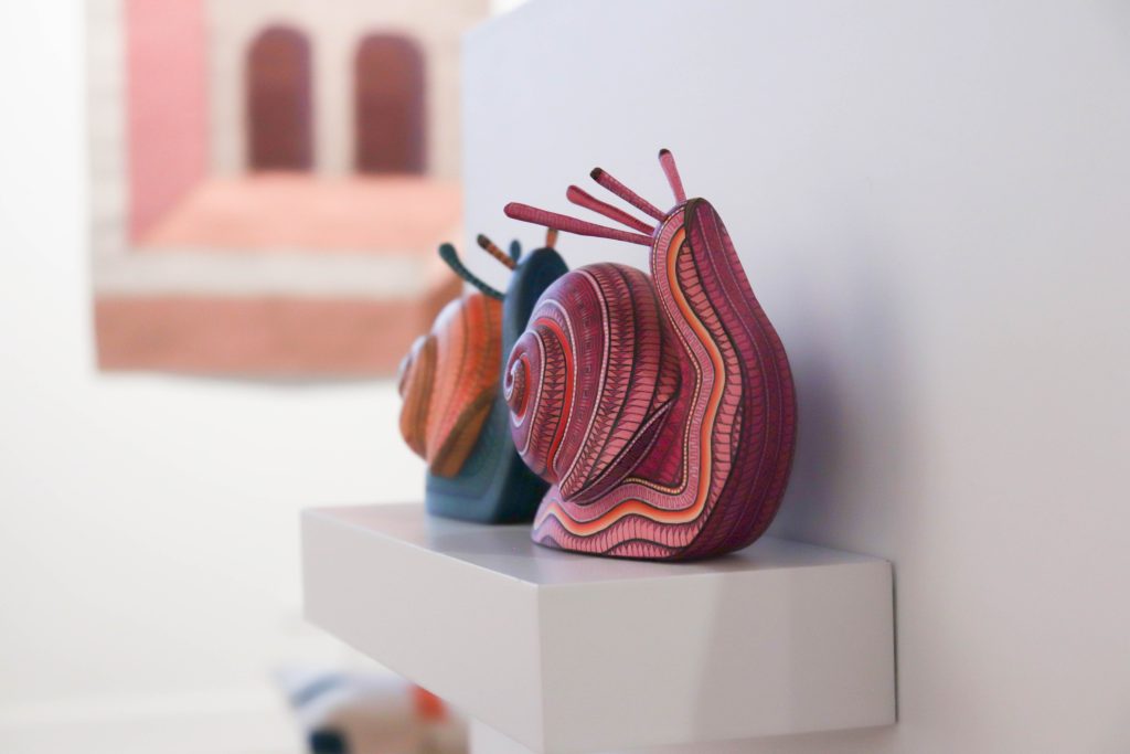 Image: Snails made by Taller Jacobo y María Ángeles on display at MANO Gallery, 2023. Two colorful snails sit on a white shelf against a white wall. Photo by Livy Snyder.