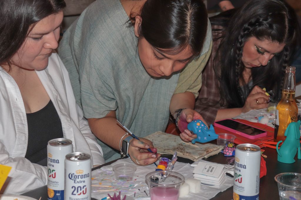 Image: María Sabina Ángeles Mendoza helping participants of Náhuatl Painting workshop a Friday evening at MANO Gallery, 2023. María leans over a table filled with art supplies with a paint brush in her hand. Photo by Livy Snyder.